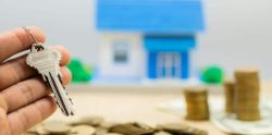 Mano sujetando una llave de vivienda, con al fondo una maqueta de casa y montones de monedas, simbolizando un impago de alquiler
