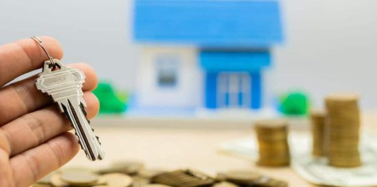 Mano sujetando una llave de vivienda, con al fondo una maqueta de casa y montones de monedas, simbolizando un impago de alquiler